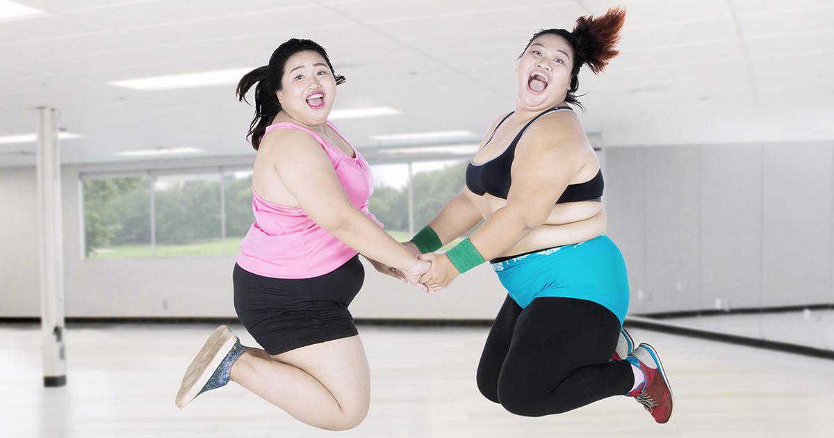 Two obese women leaping together in the gym while looking at the camera