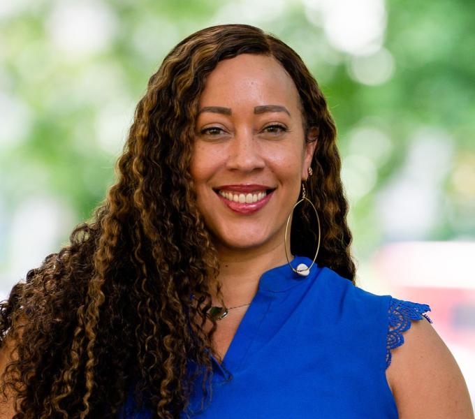 Professional headshot of a smiling woman with curly brown hair wearing a blue top and hoop earrings against a blurred green background