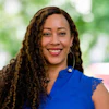 Professional headshot of a smiling woman with curly brown hair wearing a blue top and hoop earrings against a blurred green background
