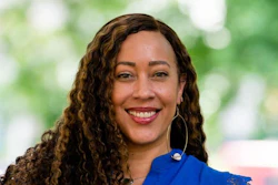 Professional headshot of a smiling woman with curly brown hair wearing a blue top and hoop earrings against a blurred green background