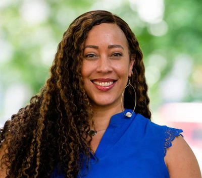 Professional headshot of a smiling woman with curly brown hair wearing a blue top and hoop earrings against a blurred green background