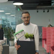 Professional man holding clipboard and plant in modern office with colleague at desk