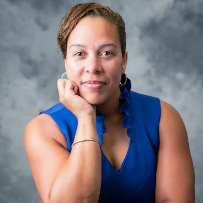 Professional headshot of a woman in a blue sleeveless top against a gray background