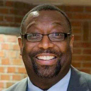 Professional headshot of a man wearing glasses and blue shirt against brick background