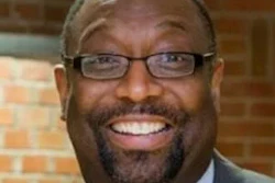 Professional headshot of a man wearing glasses and blue shirt against brick background
