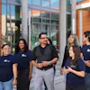 Group of six diverse professionals wearing matching navy branded t-shirts standing together outside a modern building with orange and concrete architecture