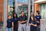 Group of six diverse professionals wearing matching navy branded t-shirts standing together outside a modern building with orange and concrete architecture