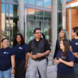 Group of six diverse professionals wearing matching navy branded t-shirts standing together outside a modern building with orange and concrete architecture