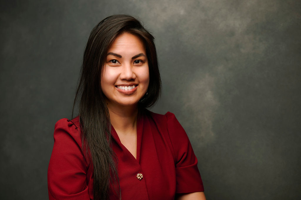 Professional headshot of smiling woman with long dark hair wearing red blazer against gray background