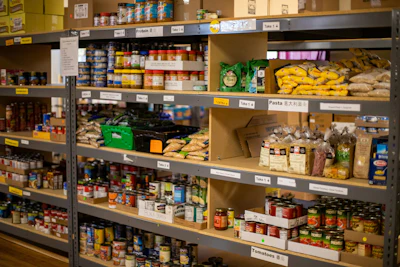 Food pantry shelves stocked with canned goods, pasta, potatoes, and packaged items organized by category