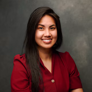 Professional headshot of a smiling woman with long dark hair wearing a burgundy blazer against a gray background