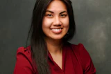 Professional headshot of a smiling woman with long dark hair wearing a burgundy blazer against a gray background