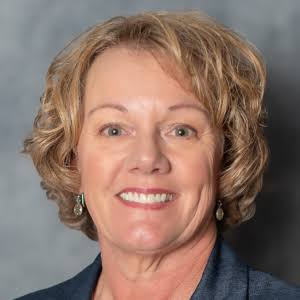 Professional headshot of a smiling woman with short blonde hair wearing a dark blue blazer and pearl earrings