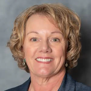 Professional headshot of a smiling woman with short blonde hair wearing a dark blue blazer and pearl earrings