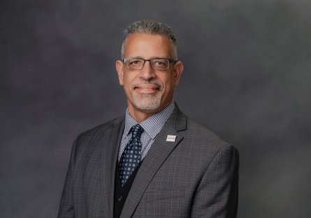 Professional headshot of a man in gray suit and blue tie wearing glasses against gray background