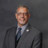 Professional headshot of a man in gray suit and blue tie wearing glasses against gray background