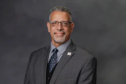 Professional headshot of a man in gray suit and blue tie wearing glasses against gray background