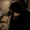 Person hunched over desk working late at night with laptop and monitor in dimly lit workspace