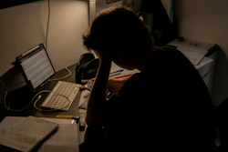 Person hunched over desk working late at night with laptop and monitor in dimly lit workspace
