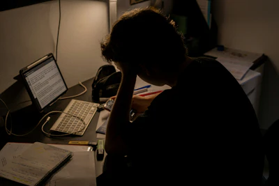 Person hunched over desk working late at night with laptop and monitor in dimly lit workspace