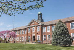Historic red brick institutional building with green cupola and manicured lawn on clear spring day