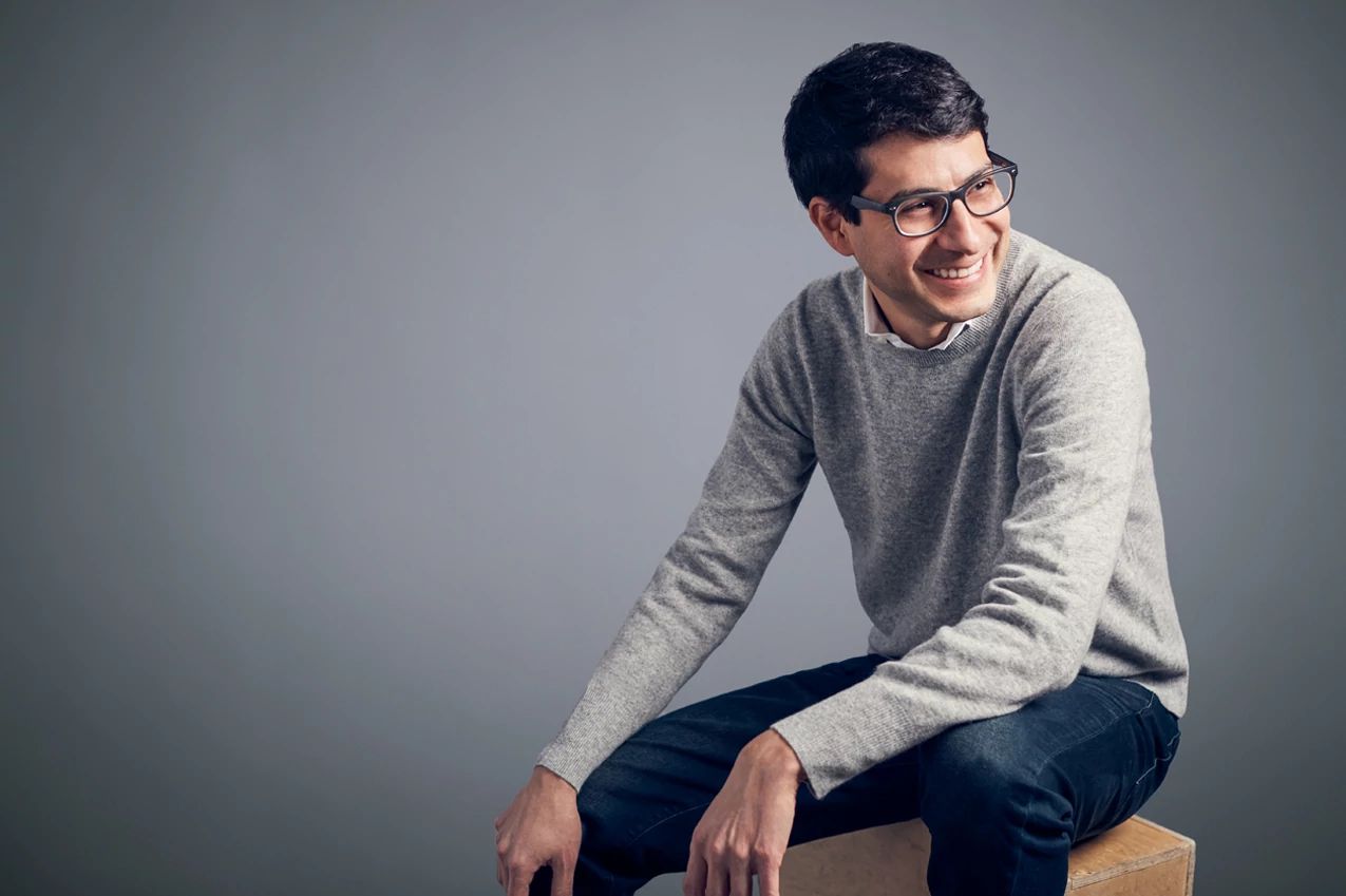 Young man wearing glasses and gray sweater sitting on wooden cube against neutral background