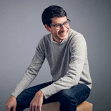 Young man wearing glasses and gray sweater sitting on wooden cube against neutral background
