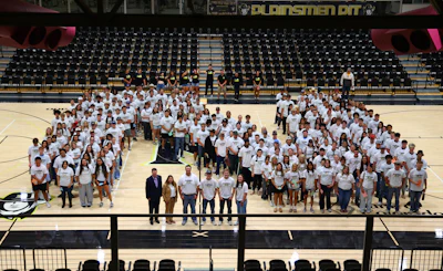Large group of students in white shirts pose for photo on gymnasium court with stadium seating in background