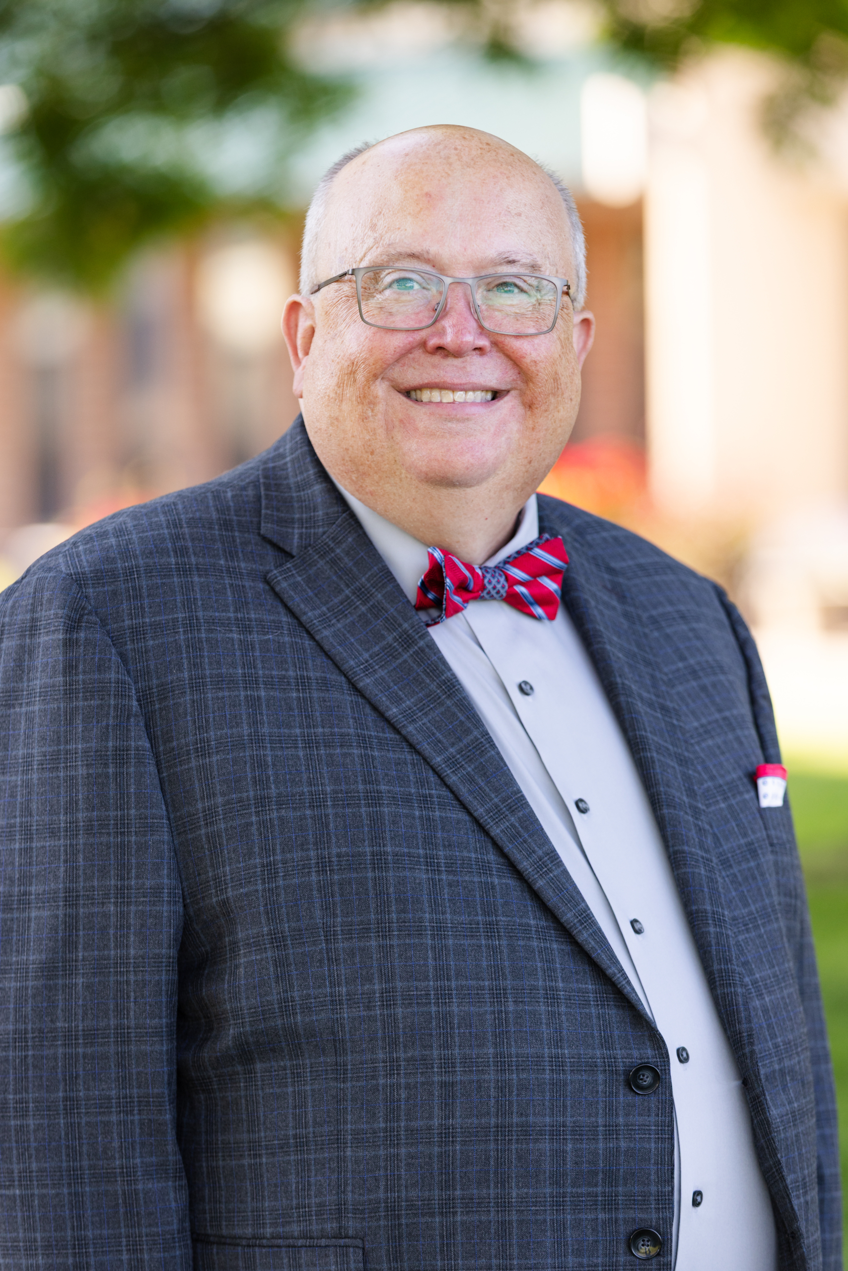 Professional headshot of a smiling man wearing glasses, navy plaid suit, light blue shirt, and red plaid bow tie