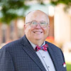 Professional headshot of a smiling man wearing glasses, navy plaid suit, light blue shirt, and red plaid bow tie