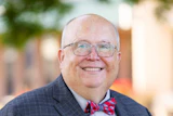 Professional headshot of a smiling man wearing glasses, navy plaid suit, light blue shirt, and red plaid bow tie