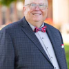 Professional headshot of a smiling man wearing glasses, navy plaid suit, light blue shirt, and red plaid bow tie