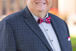 Professional headshot of a smiling man wearing glasses, navy plaid suit, light blue shirt, and red plaid bow tie