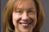 Professional headshot of a smiling woman with reddish-brown hair against a gray background