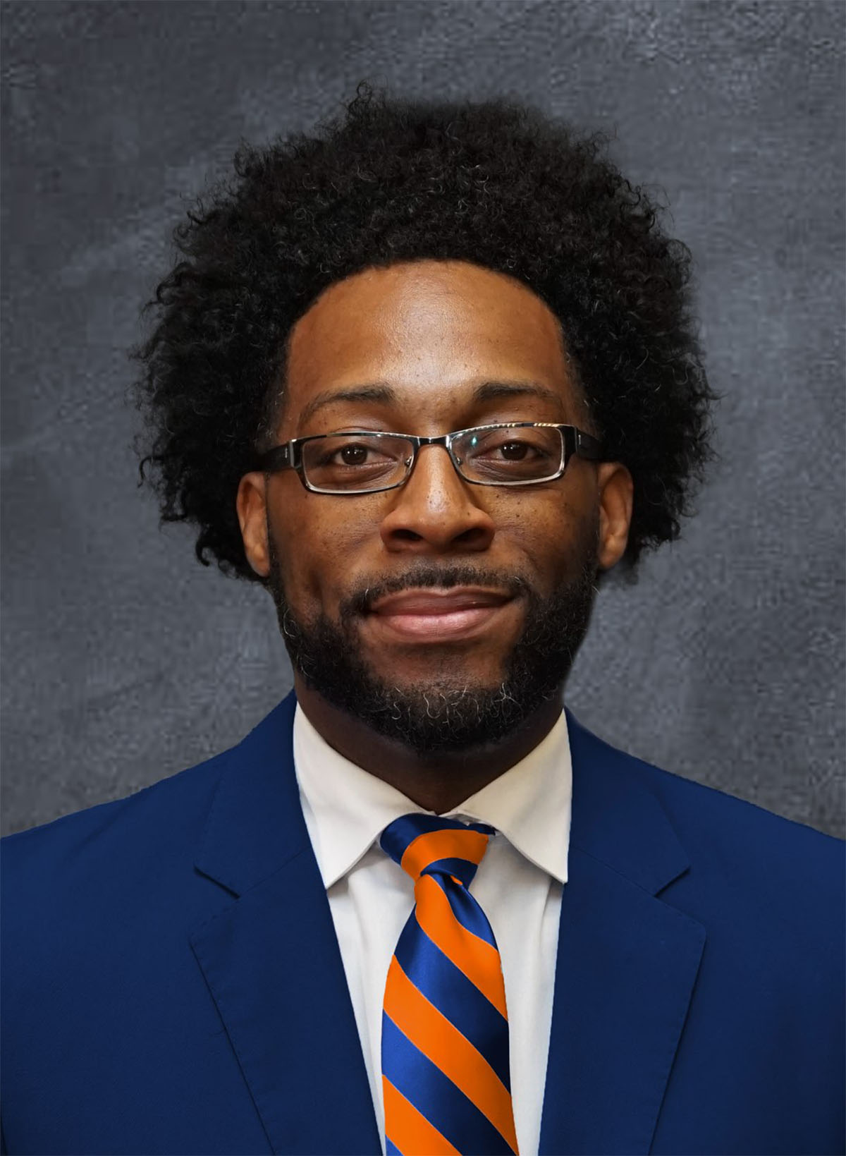 Professional headshot of a man wearing glasses, dark blue blazer, white shirt, and orange-blue striped tie against gray background