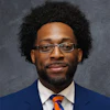 Professional headshot of a man wearing glasses, dark blue blazer, white shirt, and orange-blue striped tie against gray background