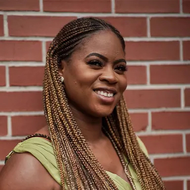 Smiling woman with braided hair wearing a green sleeveless top against a red brick wall Headshot