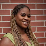 Smiling woman with braided hair wearing a green sleeveless top against a red brick wall