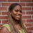 Smiling woman with braided hair wearing a green sleeveless top against a red brick wall