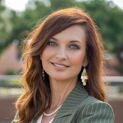 Professional portrait of a woman with reddish-brown wavy hair, pearl earrings, and pinstriped blazer smiling outdoors