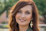 Professional portrait of a woman with reddish-brown wavy hair, pearl earrings, and pinstriped blazer smiling outdoors