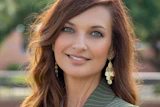 Professional portrait of a woman with reddish-brown wavy hair, pearl earrings, and pinstriped blazer smiling outdoors