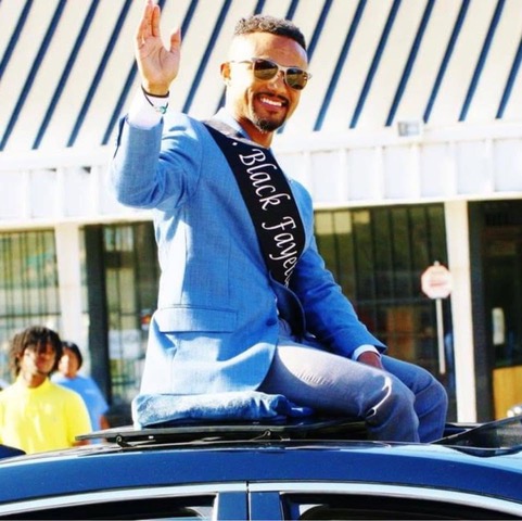 Man in blue blazer and sash waving from vehicle at outdoor event