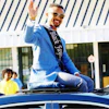 Man in blue blazer and sash waving from vehicle at outdoor event