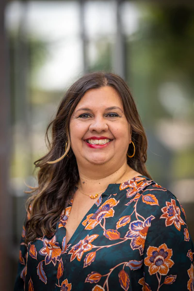 Professional headshot of a smiling woman wearing a floral patterned blouse with gold hoop earrings