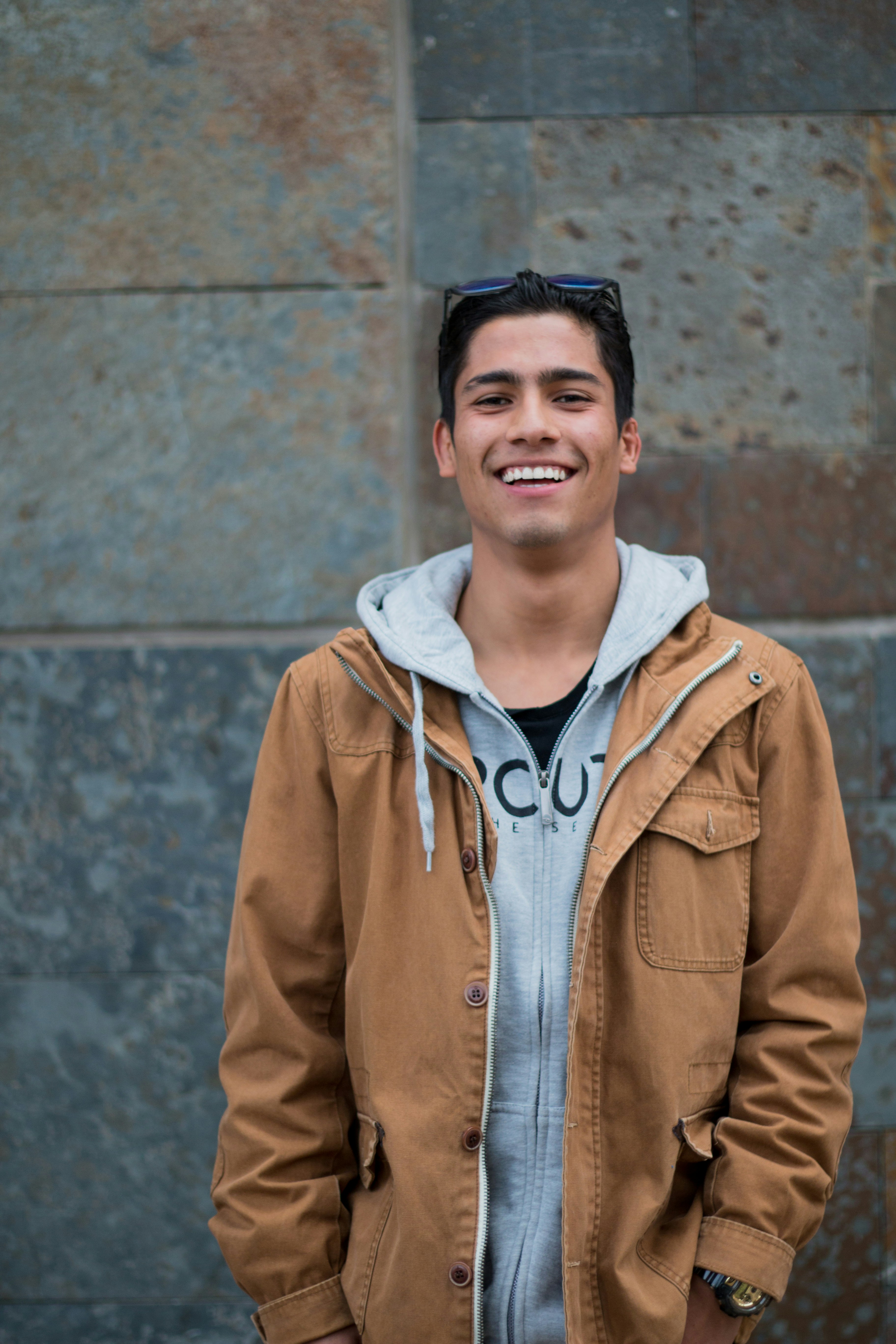 Young man smiling at camera wearing tan jacket and light blue hoodie against textured wall