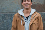 Young man smiling at camera wearing tan jacket and light blue hoodie against textured wall