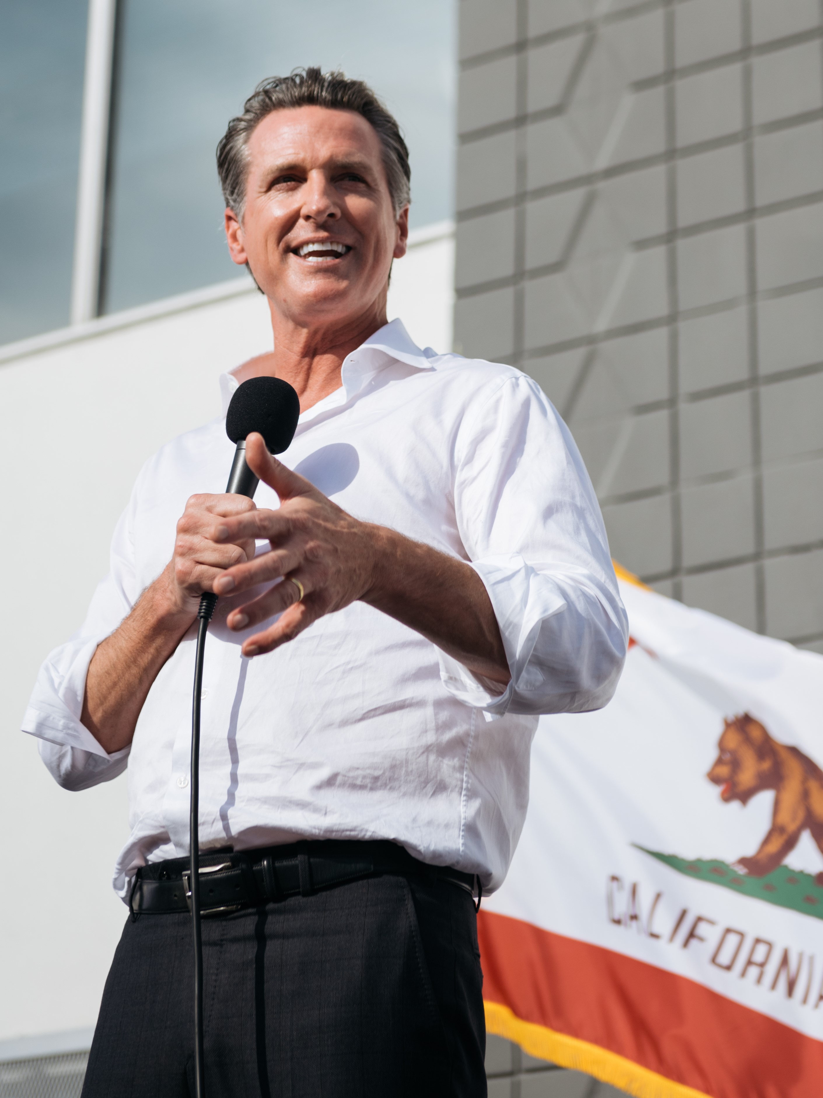 Man in white shirt speaking at microphone with California flag in background