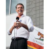 Man in white shirt speaking at microphone with California flag in background