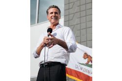 Man in white shirt speaking at microphone with California flag in background
